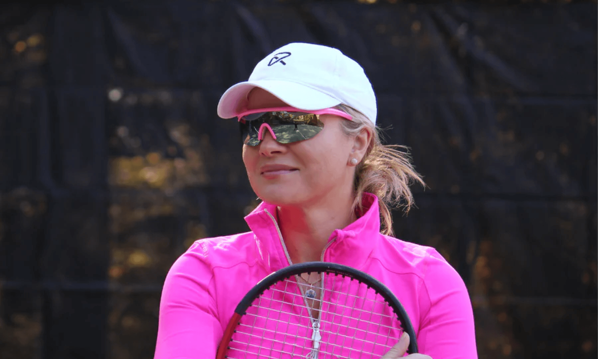 Woman in pink jacket with tennis racket, white hat and pink polarized sunglasses